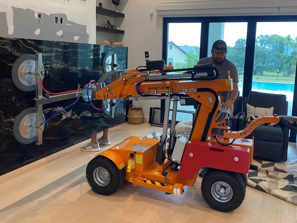 A Smartlift SL609 holds a dark marble slab flat against a living room feature wall while one person steadies it and another works the remote